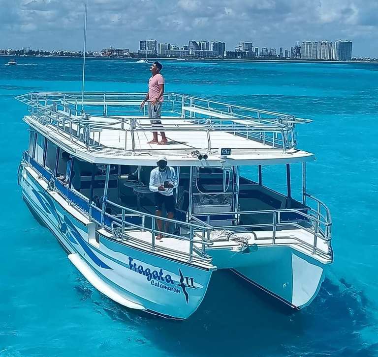 Aerial view of the white and blue Fragata II catamaran with a guide on the upper deck, anchored in turquoise Caribbean water with a city skyline in the background.