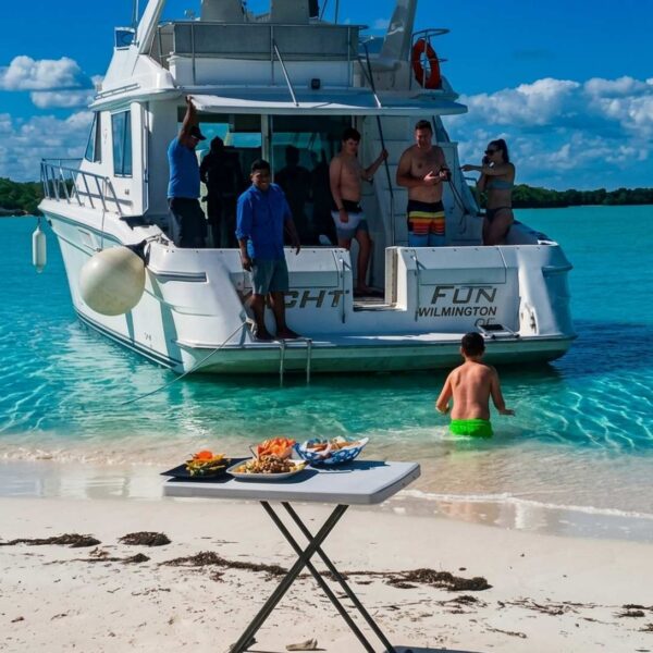 Mesa blanca montada con platones de mariscos en una playa de Canc&uacute;n junto a un yate anclado en aguas turquesas cristalinas.
