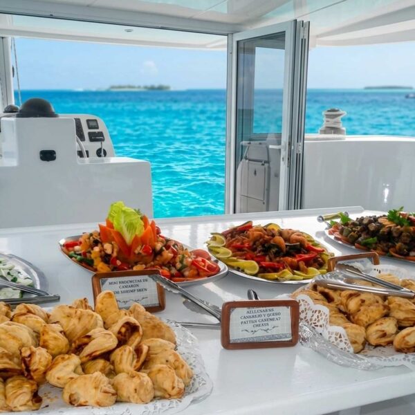Buffet en catamar&aacute;n con empanadas frescas, ensalada de camar&oacute;n y platones de mariscos servido junto al tim&oacute;n sobre aguas turquesas del Caribe.