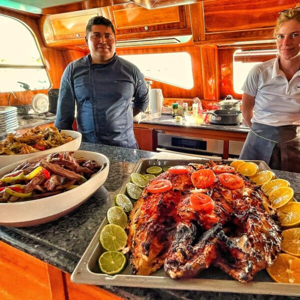 Chef privado sonriente y miembro de la tripulaci&oacute;n presentando pescado entero asado con c&iacute;tricos y fajitas en la cocina del yate.