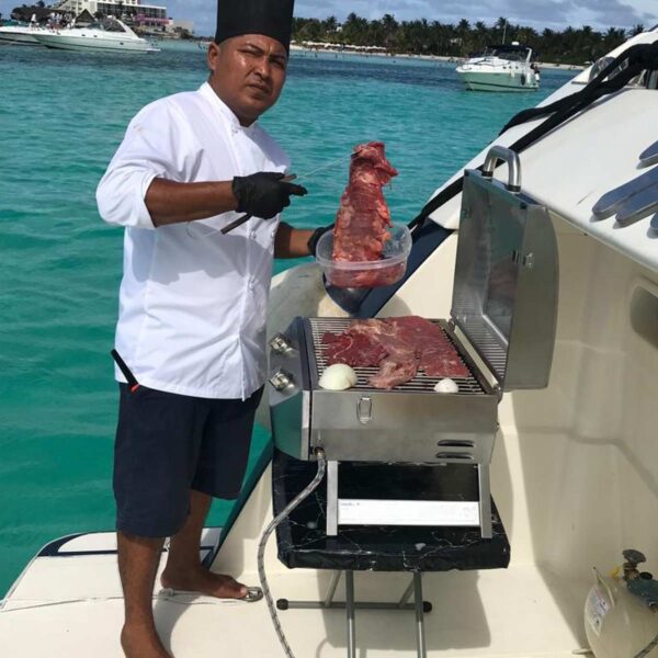 Chef con uniforme blanco asando cortes de res en la cubierta del barco con la zona hotelera de Canc&uacute;n y el mar turquesa detr&aacute;s.