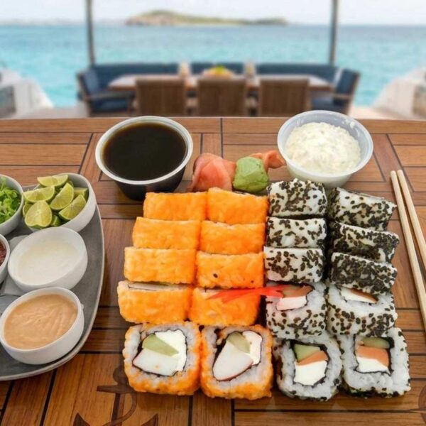 Rollos de sushi variados con salsa de soya, wasabi y toppings servidos en una mesa de teca del yate sobre aguas turquesas.