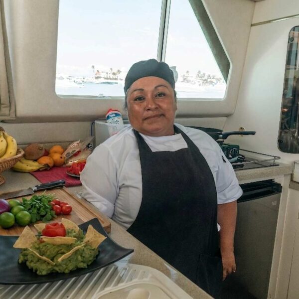 Chef sonriente en la cocina del catamar&aacute;n con una canasta de fruta fresca y un plato de guacamole con nachos.