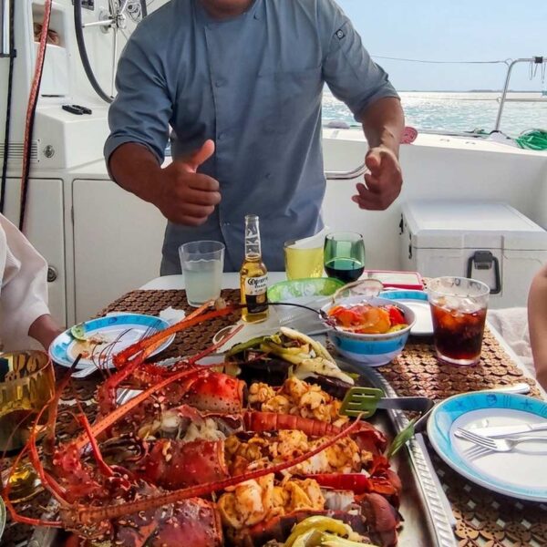 Chef privado radiante haciendo un pulgar arriba detr&aacute;s de un enorme plat&oacute;n de langosta, cortes de res y verduras a la parrilla en cubierta.