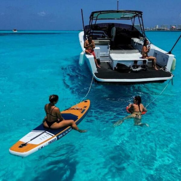 Invitado descansando sobre un paddleboard inflable junto a la plataforma de ba&ntilde;o mientras otros hacen esn&oacute;rquel al lado del yate en agua turquesa brillante.