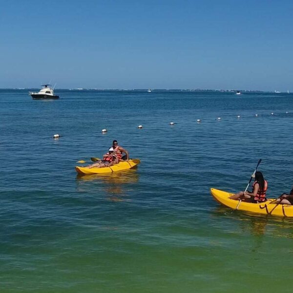 Dos kayaks tandem amarillos con invitados en chalecos salvavidas remando por la laguna calma de Canc&uacute;n cerca de un barco anclado.