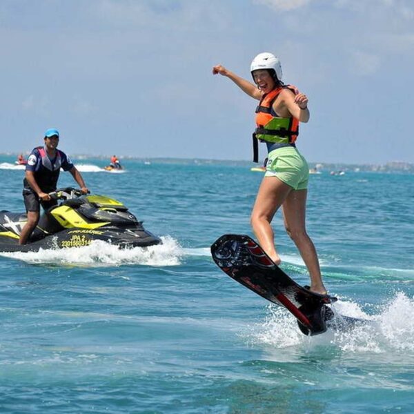 Mujer sonriente haciendo jetboard sobre una tabla con motor mientras un instructor la sigue en jet ski por aguas turquesas de Canc&uacute;n.