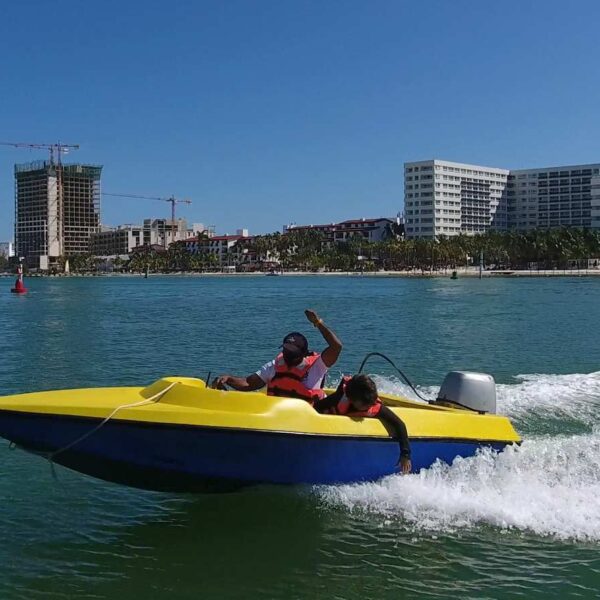 Dos personas manejan una peque&ntilde;a lancha r&aacute;pida amarilla por la laguna de Canc&uacute;n con el skyline de la zona hotelera detr&aacute;s.
