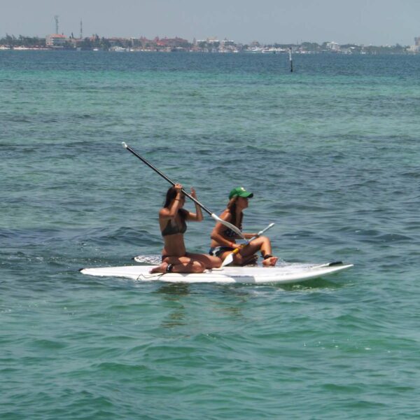 Dos mujeres comparten una tabla de stand up paddle en la laguna calma de Canc&uacute;n con la l&iacute;nea de la ciudad a lo lejos.