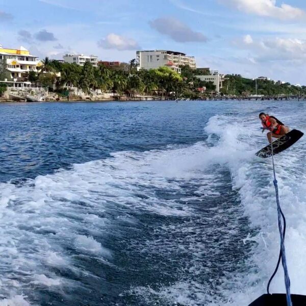 Wakeboarder con chaleco salvavidas cortando la estela del barco a lo largo de la costa de Isla Mujeres en un d&iacute;a soleado.