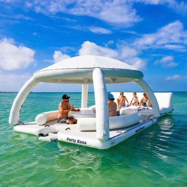 AquaBanas Party Bana shaded floating cabana anchored in turquoise Caribbean shallows with five guests lounging in the shade in the middle of the sea.