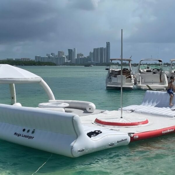 AquaBanas Mega Lounger inflatable floating dock with a shaded cabana and extended sun deck where two guests sit, anchored in calm green-blue marina water.