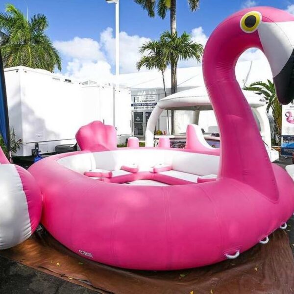 Giant pink flamingo inflatable float for up to six guests with cup holders and central cushioned seating, displayed at a Florida marine boat show.