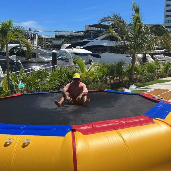 Floating inflatable trampoline with bright yellow base and colourful trim at the Cancun marina with palms and luxury yachts in the background.